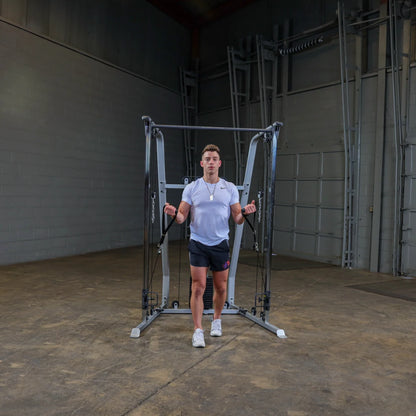 A person standing in a gym setting, using a cable machine for strength training. They are wearing a short-sleeved shirt and shorts, holding the handles of the machine with both hands. The background features gray walls and gym equipment.