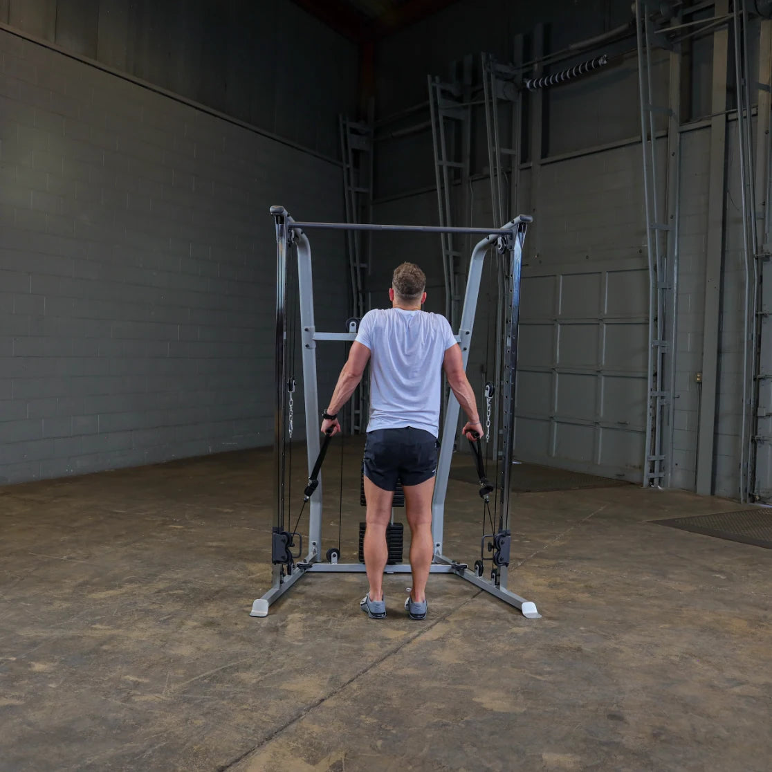 A person in a white shirt and black shorts is using a cable machine for exercise, facing away from the camera in a spacious gym with gray walls and a concrete floor.