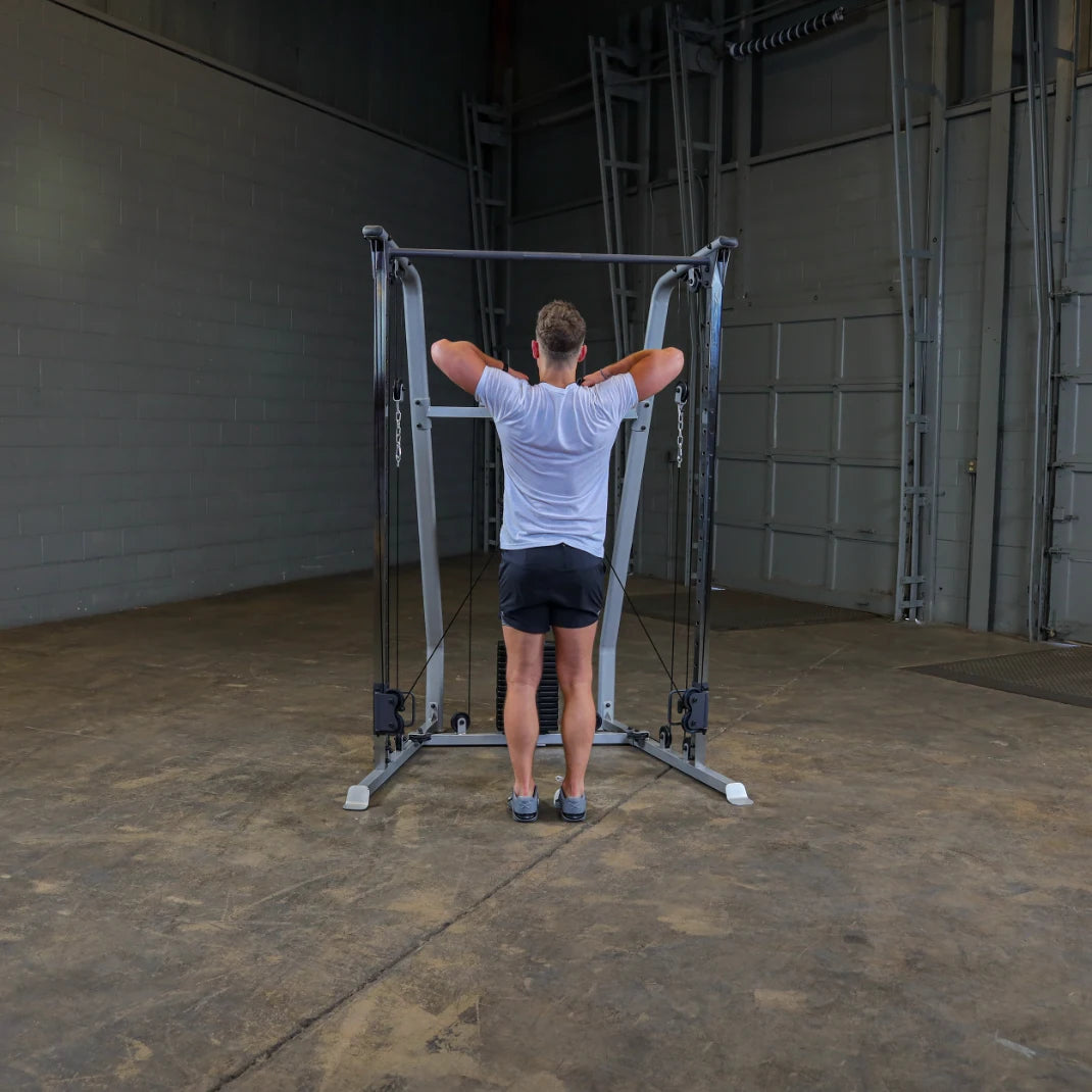 A person standing in a gym setting, performing an exercise on a cable machine, facing away from the camera, wearing a white t-shirt and black shorts.