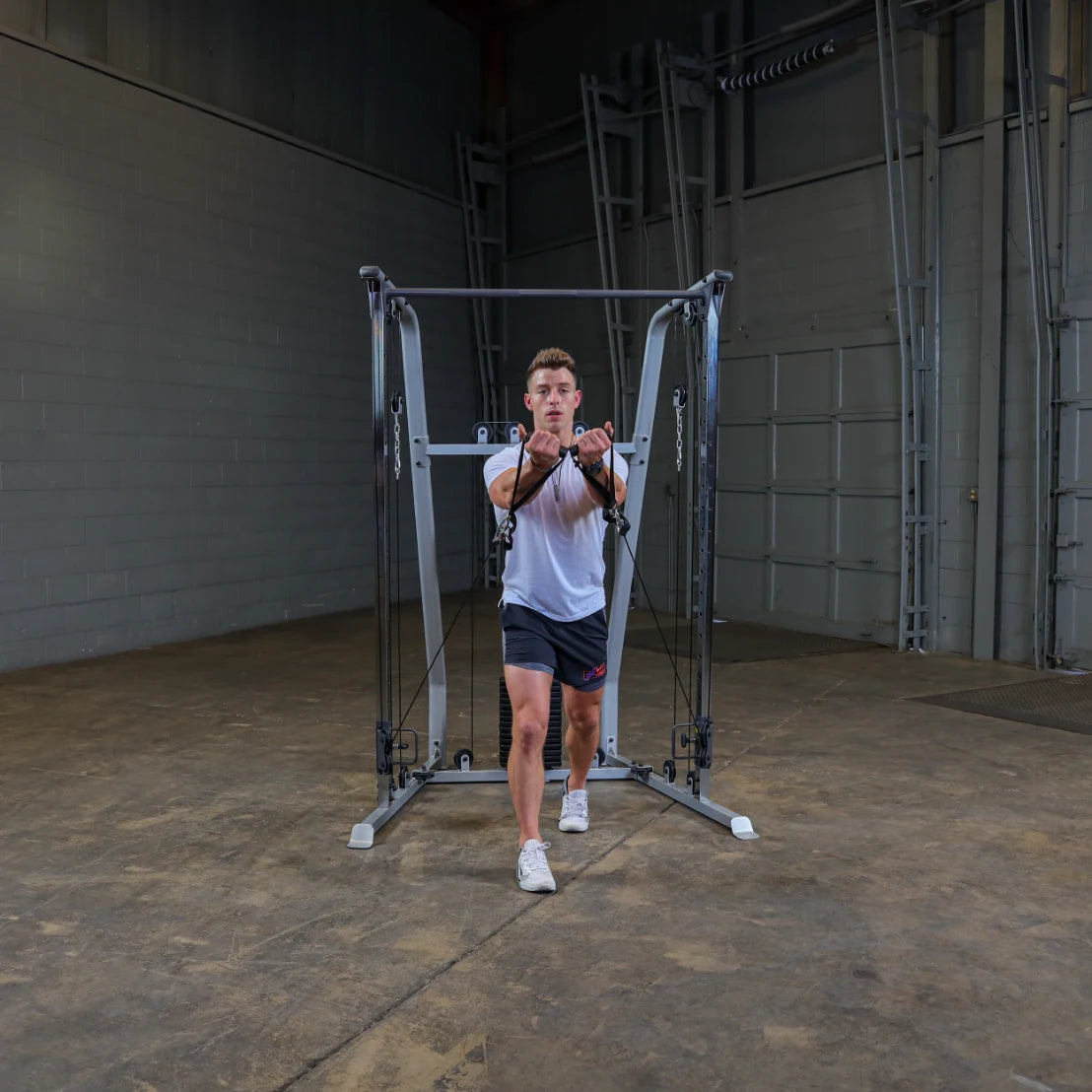 A person in a white t-shirt and black shorts performing a cable exercise in a gym setting, using a cable machine with adjustable pulleys. The background features a spacious, industrial-style gym with gray walls and minimal equipment.