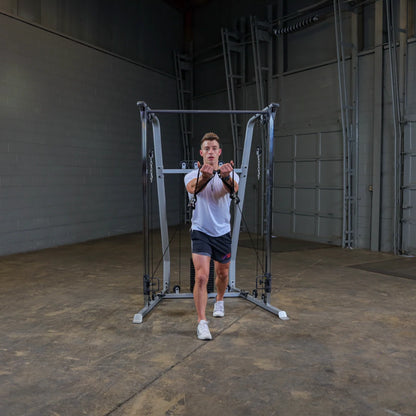 A person in a white t-shirt and black shorts performing a cable exercise in a gym setting, using a cable machine with adjustable pulleys. The background features a spacious, industrial-style gym with gray walls and minimal equipment.