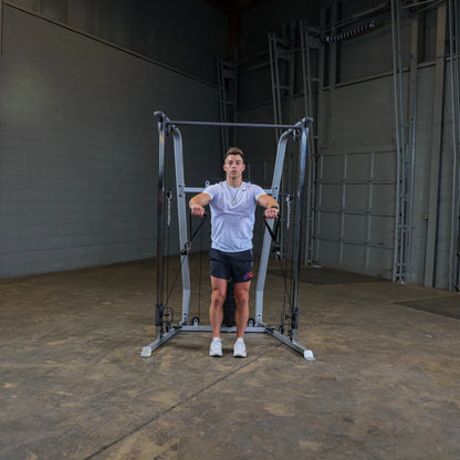 A young man stands in front of a cable machine in a gym setting, with his arms extended out to the sides, demonstrating a workout posture. The background features a gray wall and an open space.