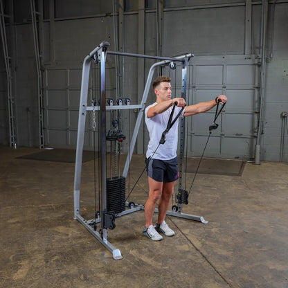 A man performing a cable exercise on a gym machine, holding the handles in front of him, in a spacious gym environment with a concrete floor and metal walls.