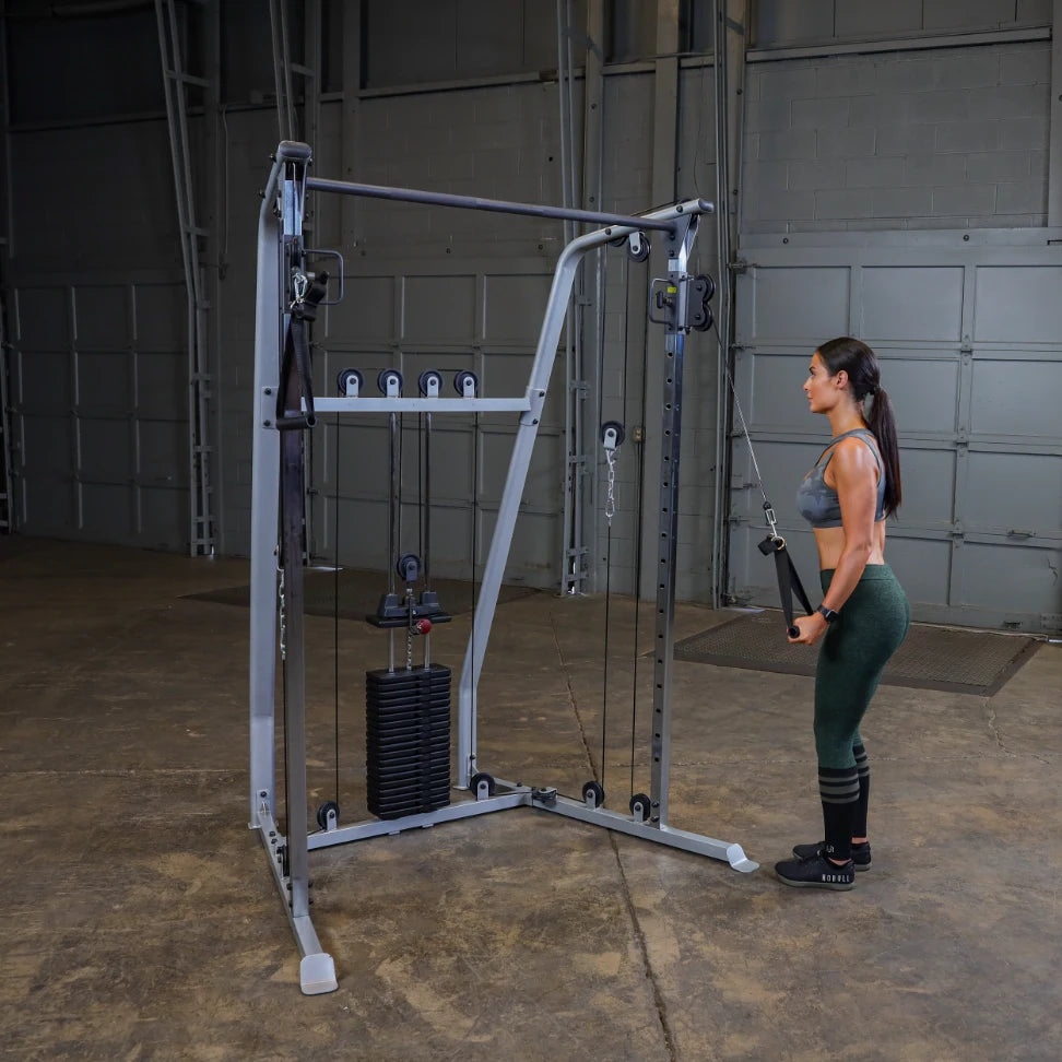 A woman with long hair is standing next to a cable machine in a gym, preparing to perform an exercise. She is wearing a gray sports bra and green leggings. The gym has a concrete floor and large garage-style doors in the background.