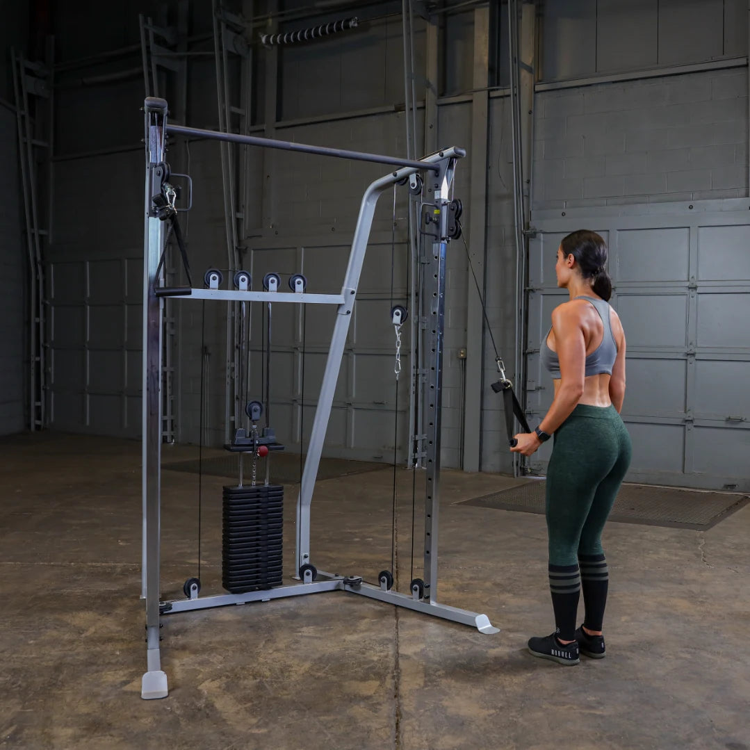 A person stands in front of a cable machine in a gym setting, preparing to perform an exercise. The machine is equipped with weights and pulleys, and the person is wearing a sports bra and leggings, focusing on their workout.