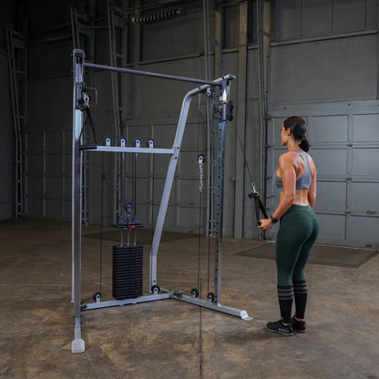 A person stands in front of a cable machine in a gym setting, preparing to perform an exercise. The machine is equipped with weights and pulleys, and the person is wearing a sports bra and leggings, focusing on their workout.
