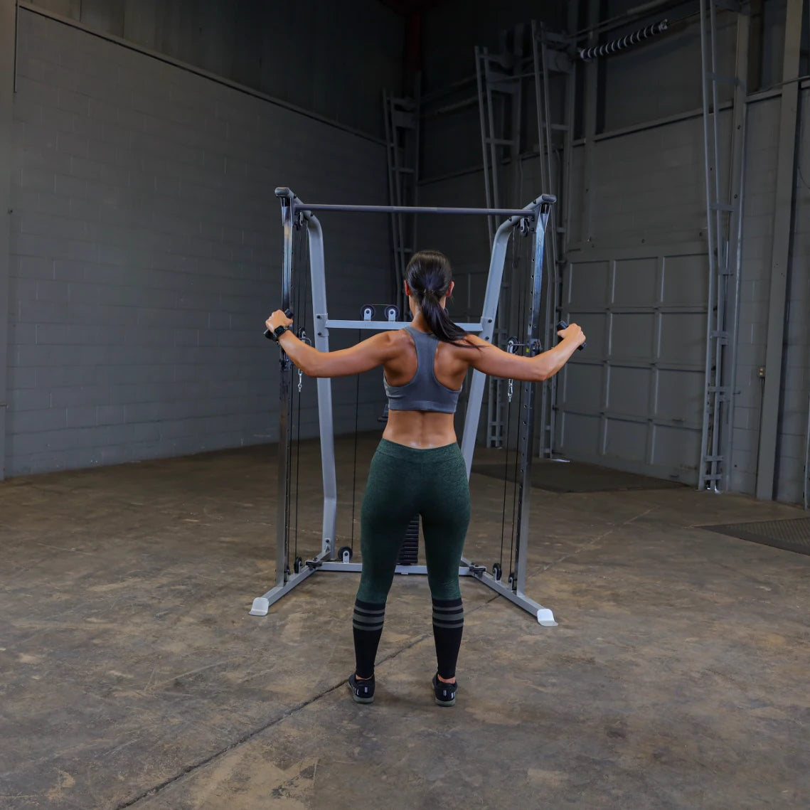 A person performing a workout on a cable machine in a gym setting, wearing a gray sports bra and green leggings, with their back facing the camera.