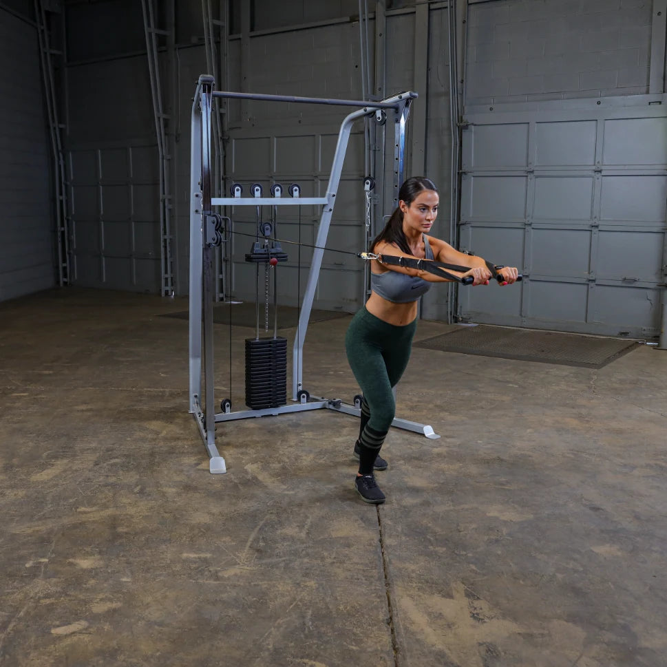 A woman performs an exercise using a cable machine in a gym setting, wearing a gray crop top and green leggings.