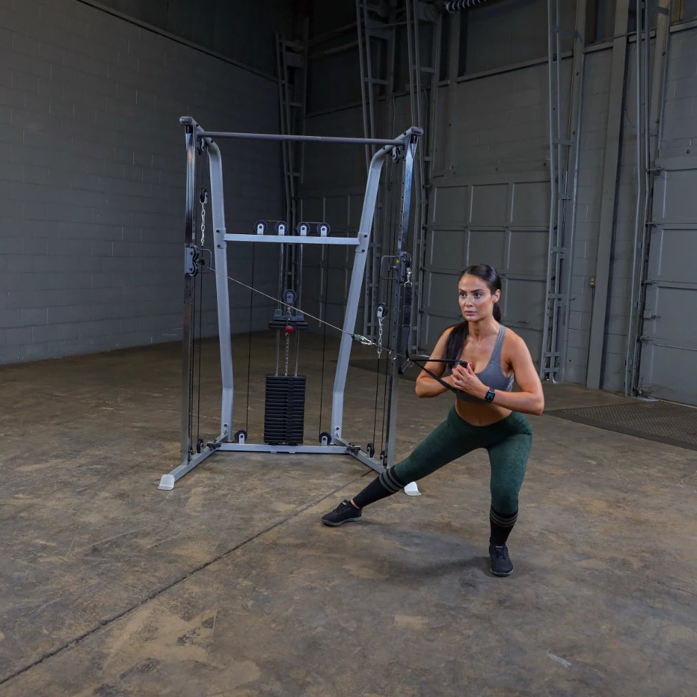 A woman performs a resistance training exercise using a cable machine in a gym setting, wearing a gray sports bra and green leggings, with a focused expression.