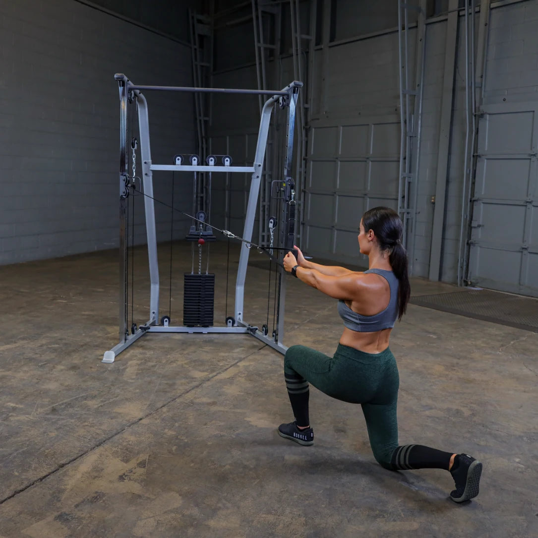 A woman performing a cable lunge exercise in a gym, using a cable machine with weights, wearing a gray sports bra and green leggings.