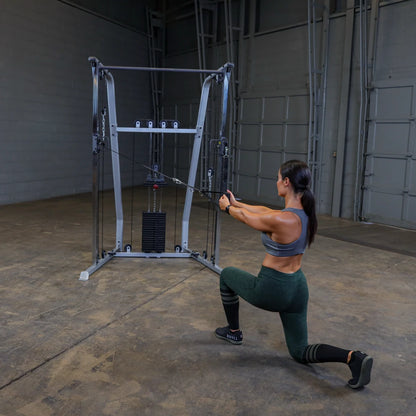 A woman performing a cable lunge exercise in a gym, using a cable machine with weights, wearing a gray sports bra and green leggings.