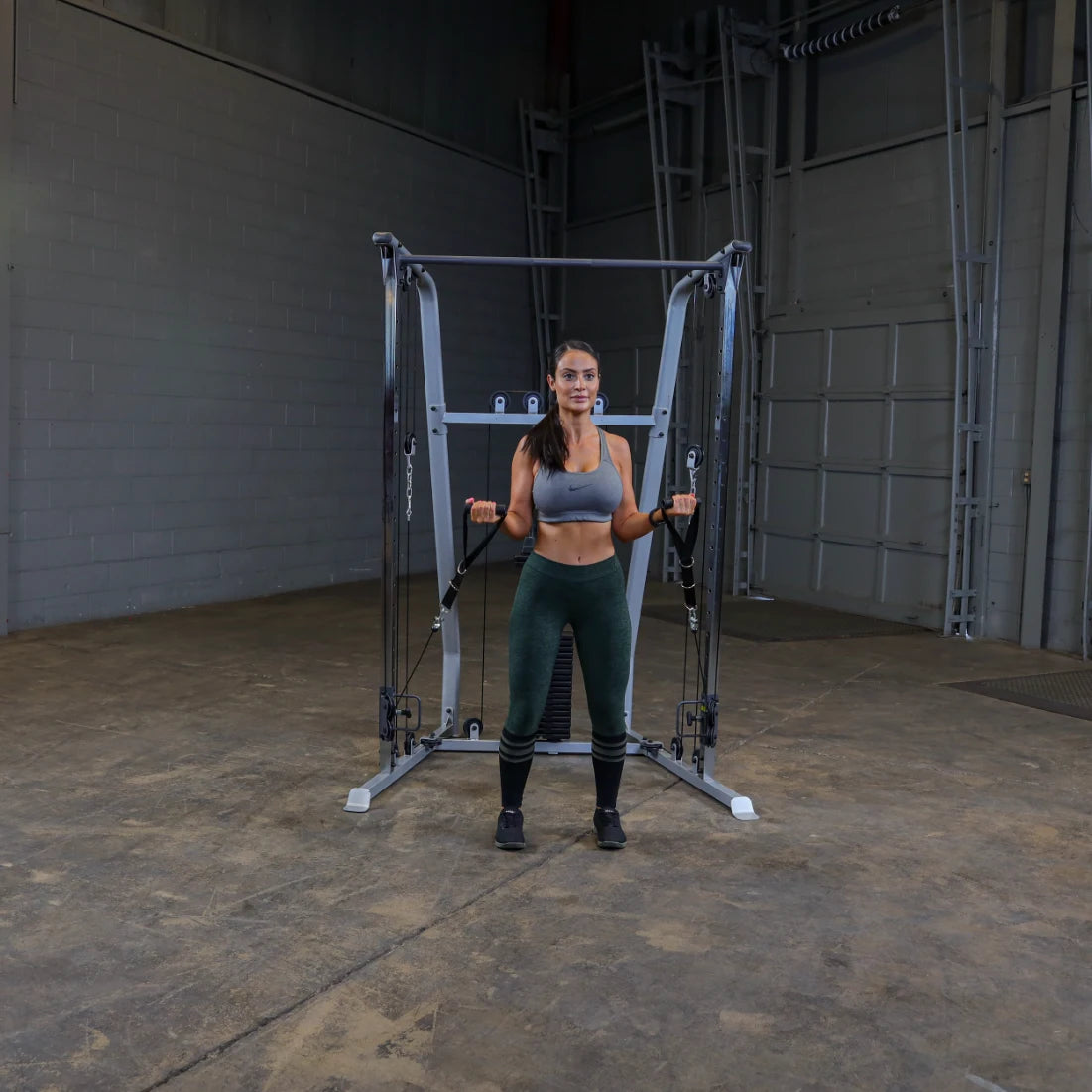 A woman in athletic wear stands in front of a cable machine in a gym, holding the handles with both hands, ready to perform an exercise. The gym has a concrete floor and a simple, industrial background.