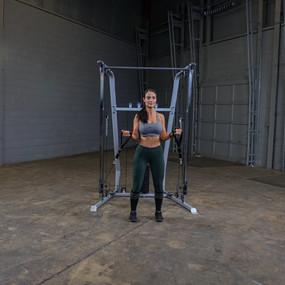 A woman in athletic wear stands in front of a cable machine in a gym, holding the handles with both hands, ready to perform an exercise. The gym has a concrete floor and a simple, industrial background.