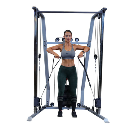 A woman in athletic wear performs a cable exercise using a gym machine, focusing on strength training. She has a serious expression and is holding the cables with both hands, positioned in front of her body.
