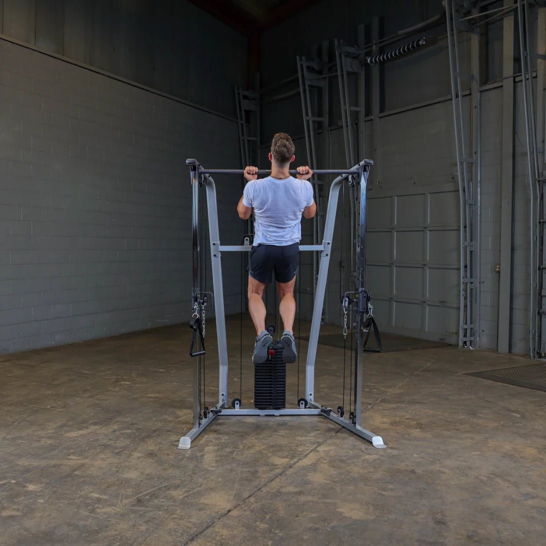 A person performing a pull-up on a gym machine in a spacious indoor setting with gray walls and a concrete floor. The individual is wearing a light gray t-shirt and black shorts.