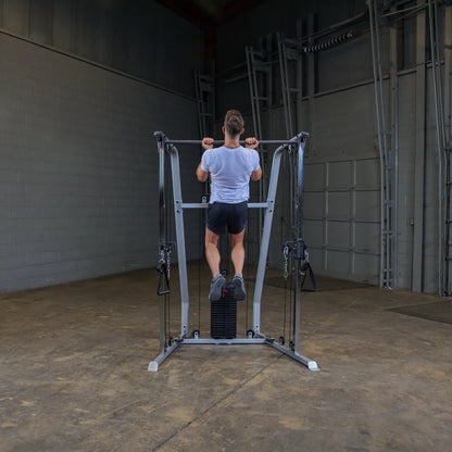 A person performing a pull-up on a gym machine in a spacious indoor setting with gray walls and a concrete floor. The individual is wearing a light gray t-shirt and black shorts.