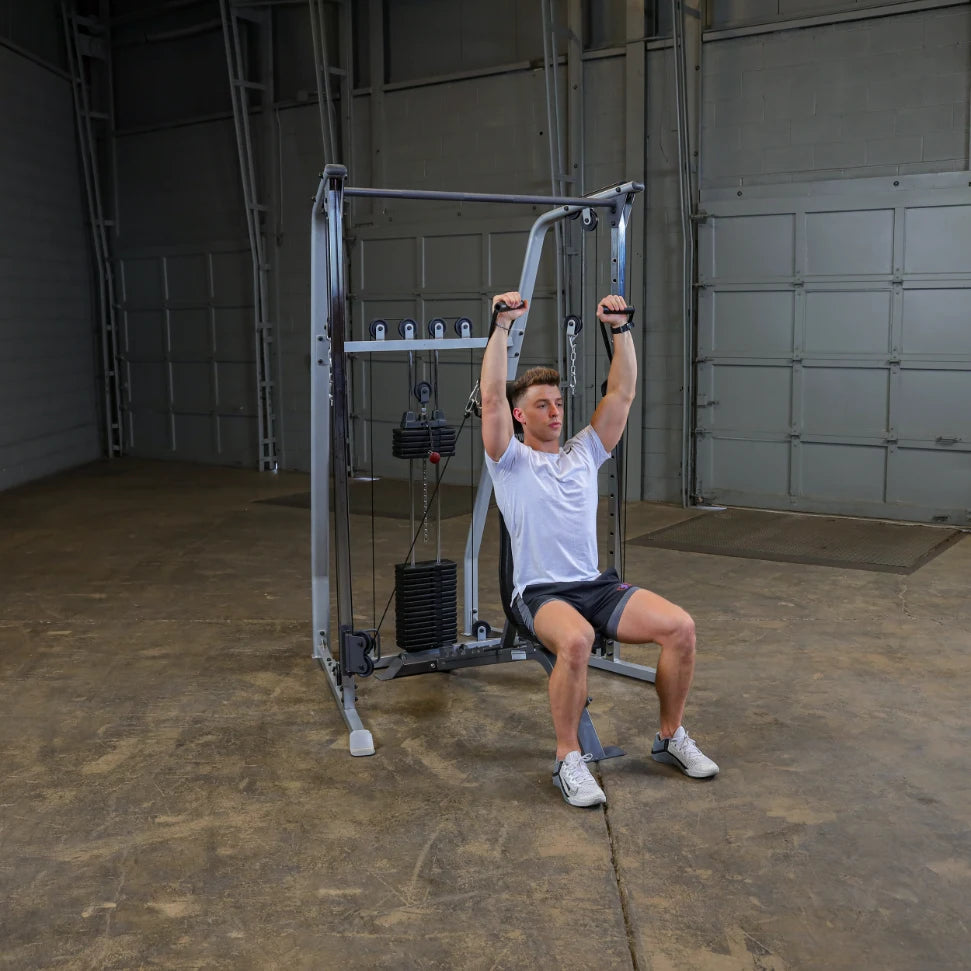 A young man performing an exercise on a cable machine in a gym setting, with a focused expression while holding onto the handles above his head.