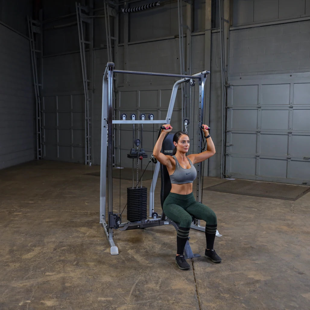 A woman performing an overhead exercise on a cable machine in a gym setting, wearing a sports bra and leggings, with a focused expression.