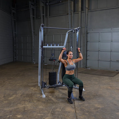 A woman performing an overhead exercise on a cable machine in a gym setting, wearing a sports bra and leggings, with a focused expression.