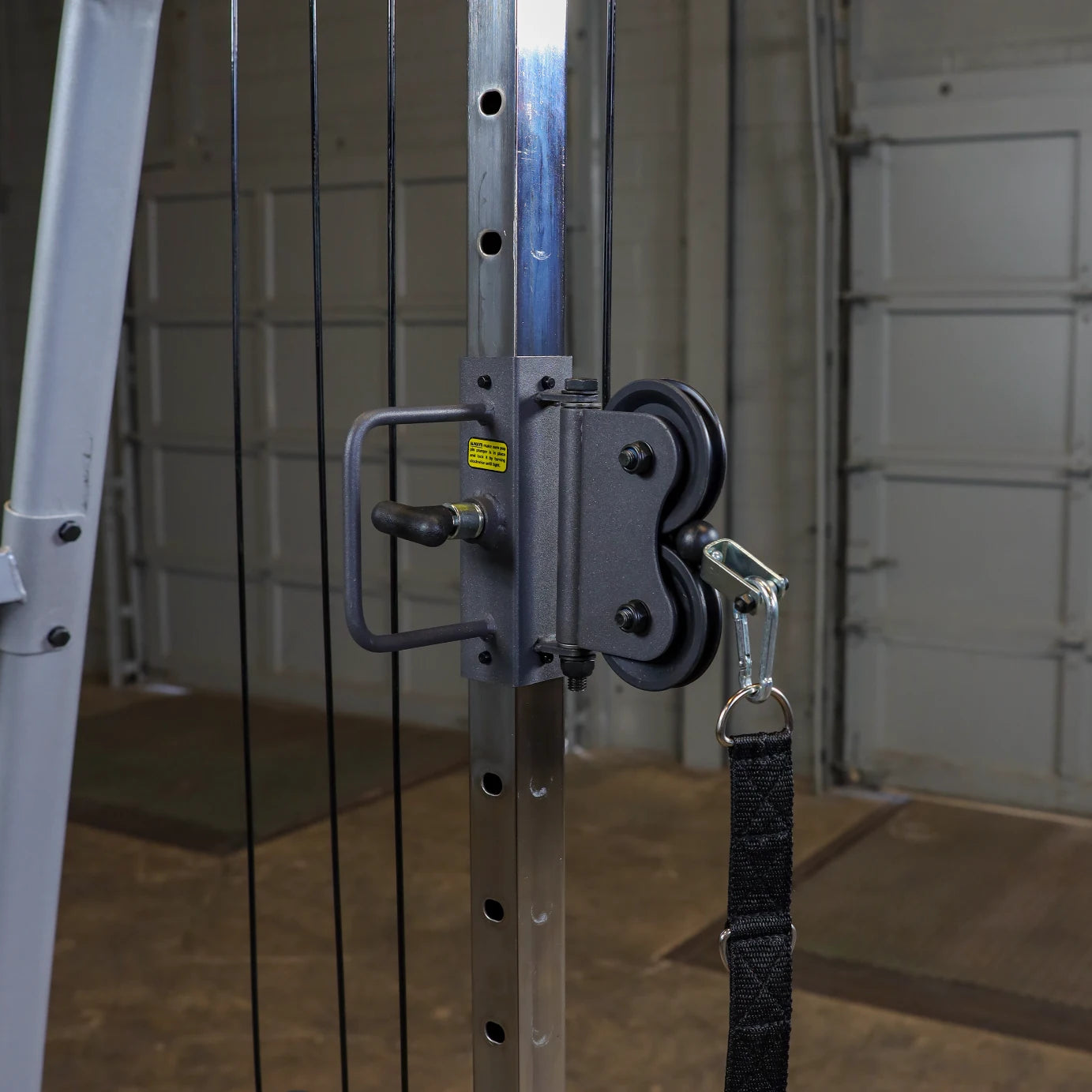 Close-up of a cable pulley system attached to a gym machine, featuring a grey metal frame, a black strap, and a carabiner, with a blurred background of a garage setting.