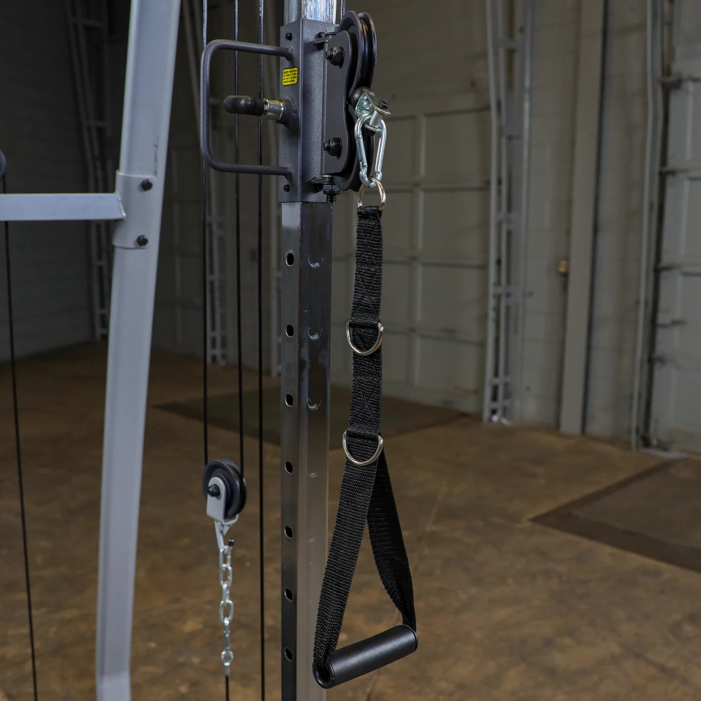 Close-up of a gym cable machine with a black strap attachment hanging next to a pulley system, set against a blurred industrial background.