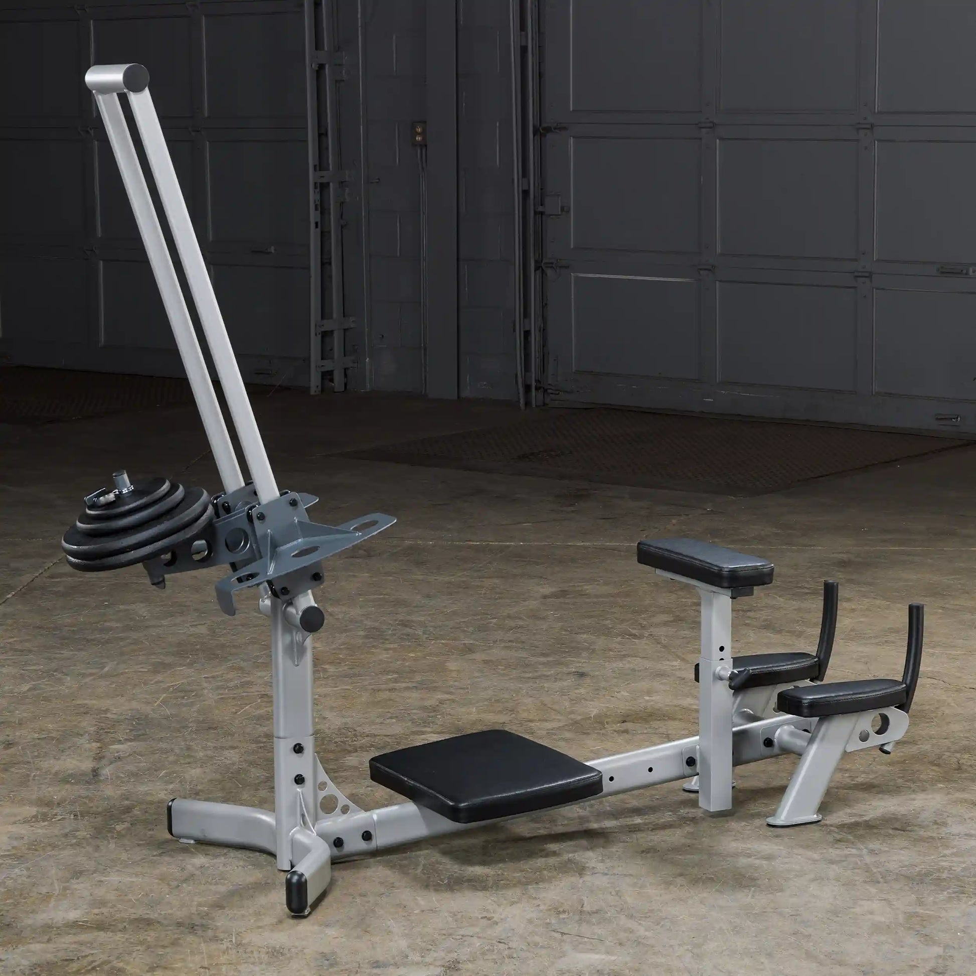 A silver and black multi-functional gym machine with a weight plate attached, set against a concrete floor and a garage door background.