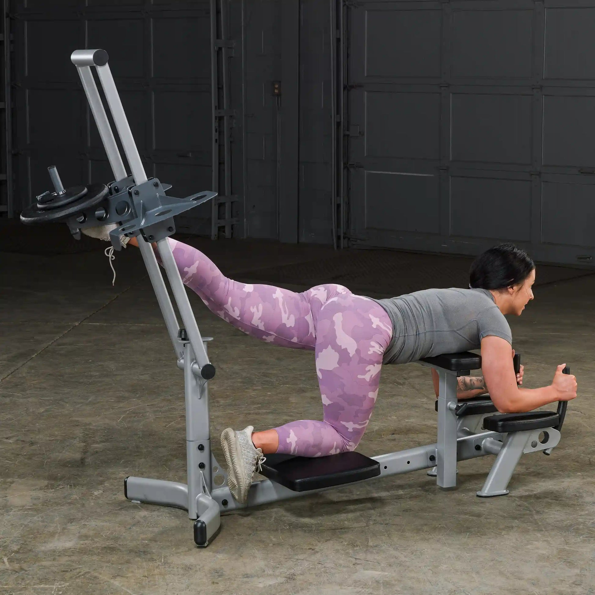 A woman in a gray workout top and pink camo leggings is performing an exercise on a gym machine, focusing on her glutes, in a spacious indoor setting.