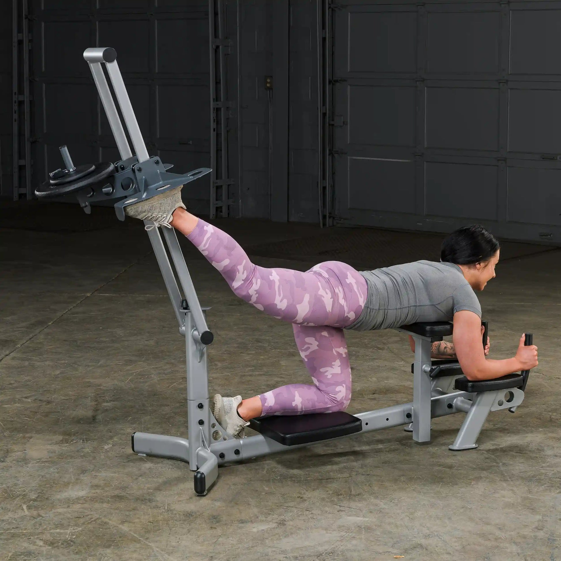 A woman in a gray athletic shirt and pink camo leggings performs an exercise on a workout bench, extending one leg backward while holding onto the equipment for support. The setting is a gym with a concrete floor and a garage-style backdrop.
