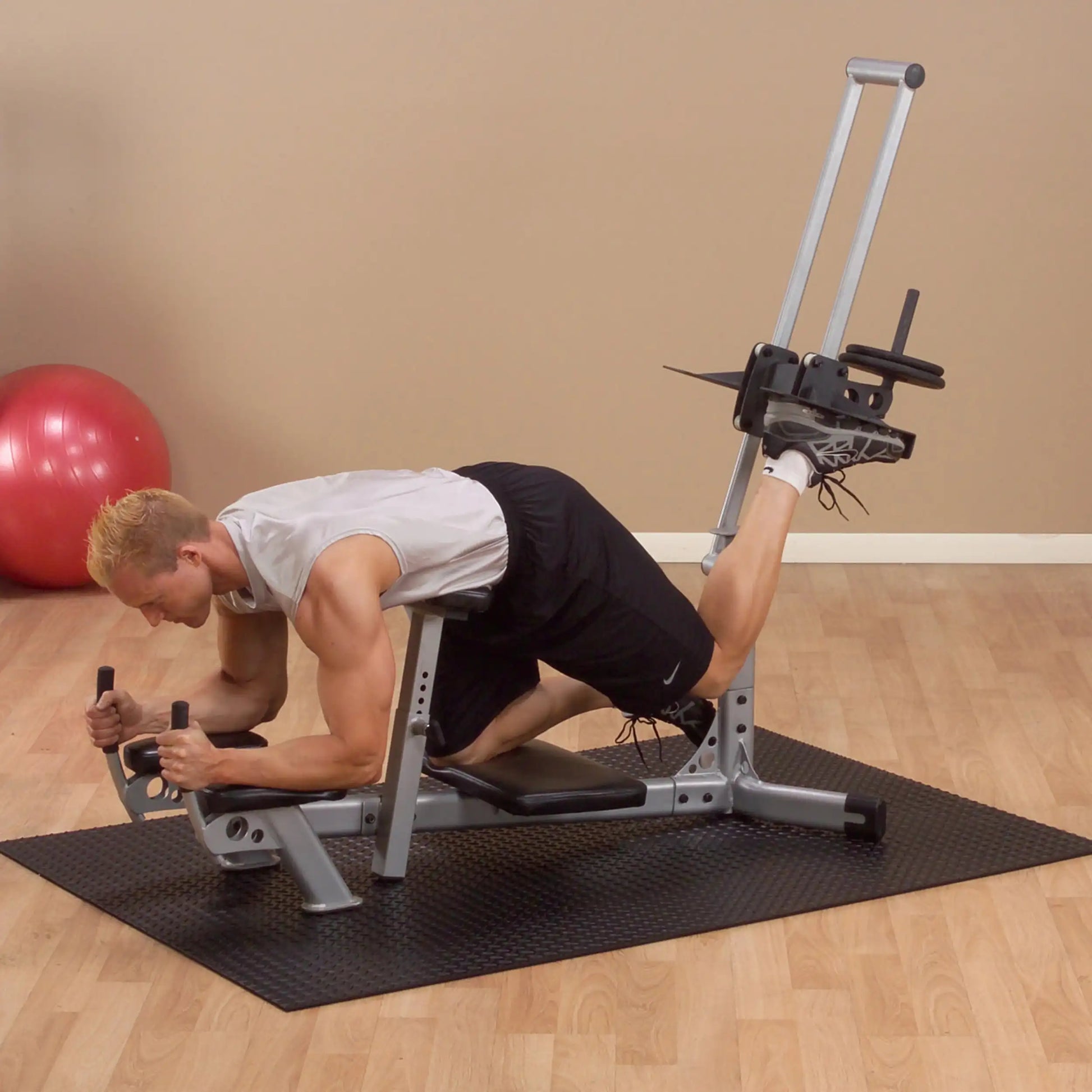 A man in athletic attire performs a leg exercise on a gym machine, focusing on strength training, with a red exercise ball in the background.