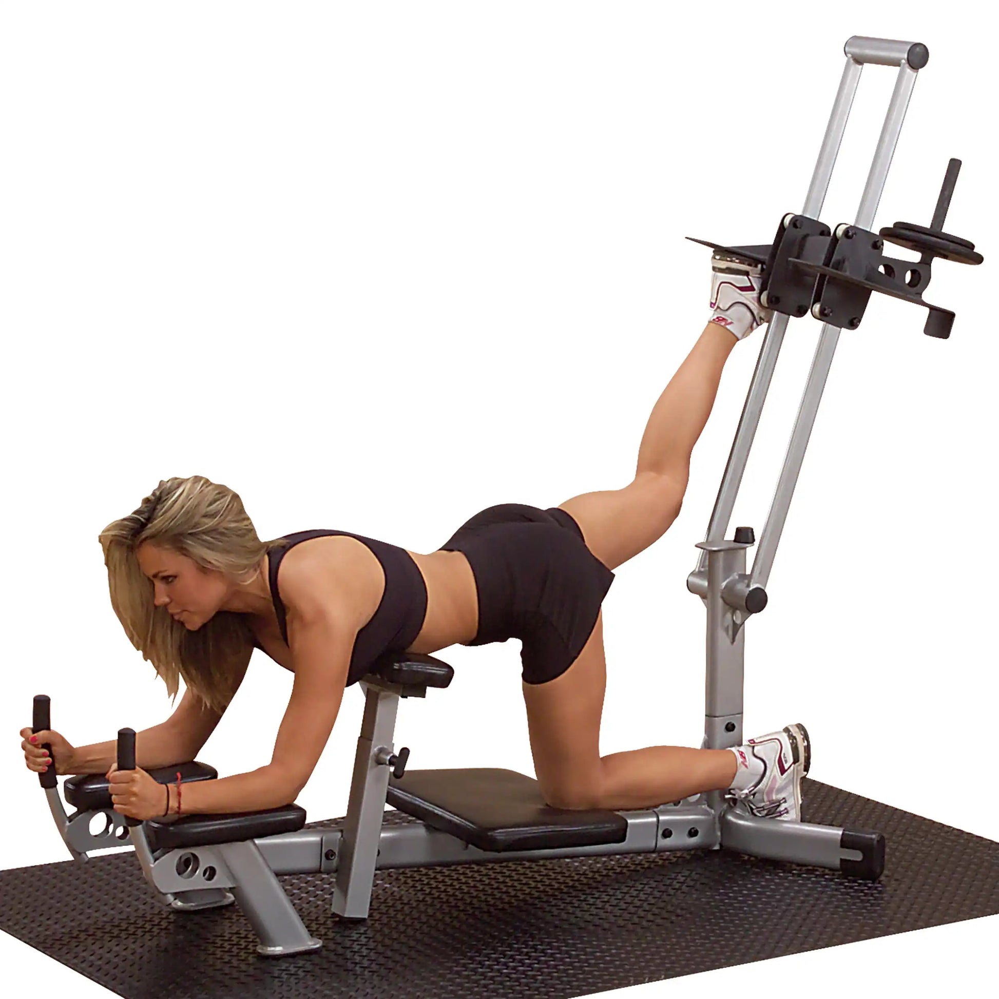 A woman performing an exercise on a gym machine, lying on her stomach with one leg extended behind her, wearing a black sports top and shorts.