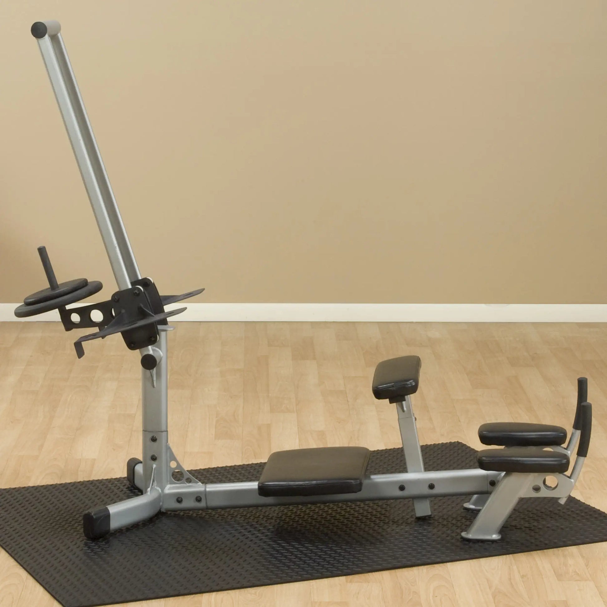 A silver weight bench with adjustable components, including a barbell holder and padded seats, placed on a black rubber mat in a room with light-colored walls.