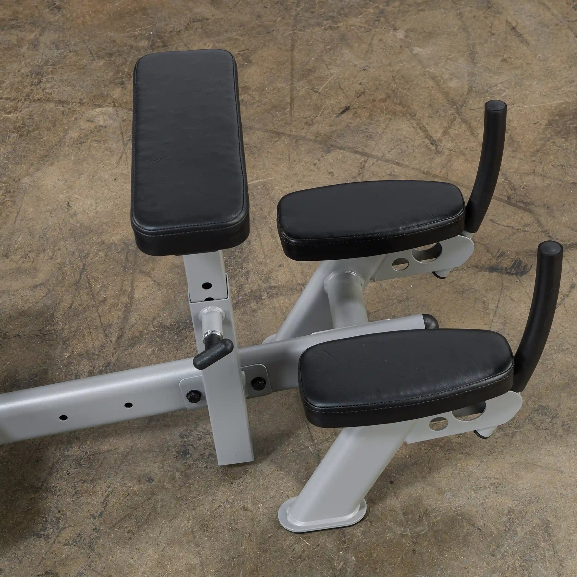 Close-up view of a piece of gym equipment featuring two black padded armrests on a silver metallic frame, set against a textured floor surface.