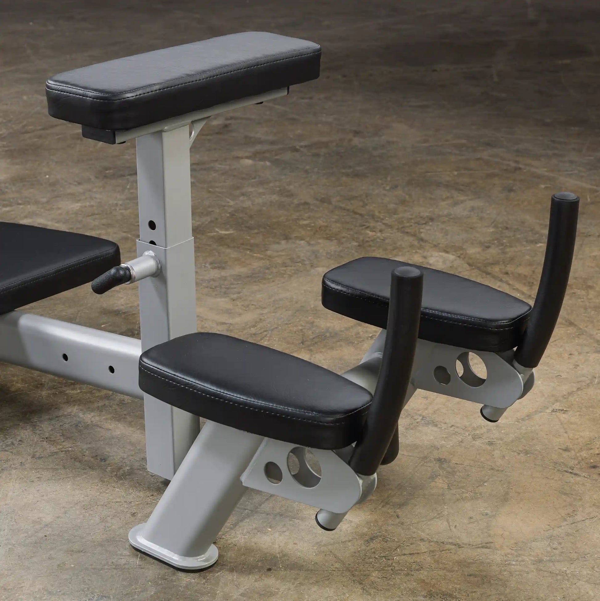 Close-up view of a weight bench with padded armrests and a back support, designed for strength training exercises. The bench features a silver metallic frame and black cushioning, set against a textured concrete floor.