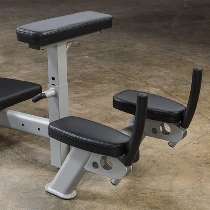 Close-up view of a weight bench with padded armrests and a back support, designed for strength training exercises. The bench features a silver metallic frame and black cushioning, set against a textured concrete floor.