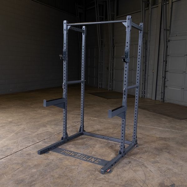 A sturdy black squat rack positioned on a concrete floor in a gym setting, with a simple backdrop of gray walls and doors.