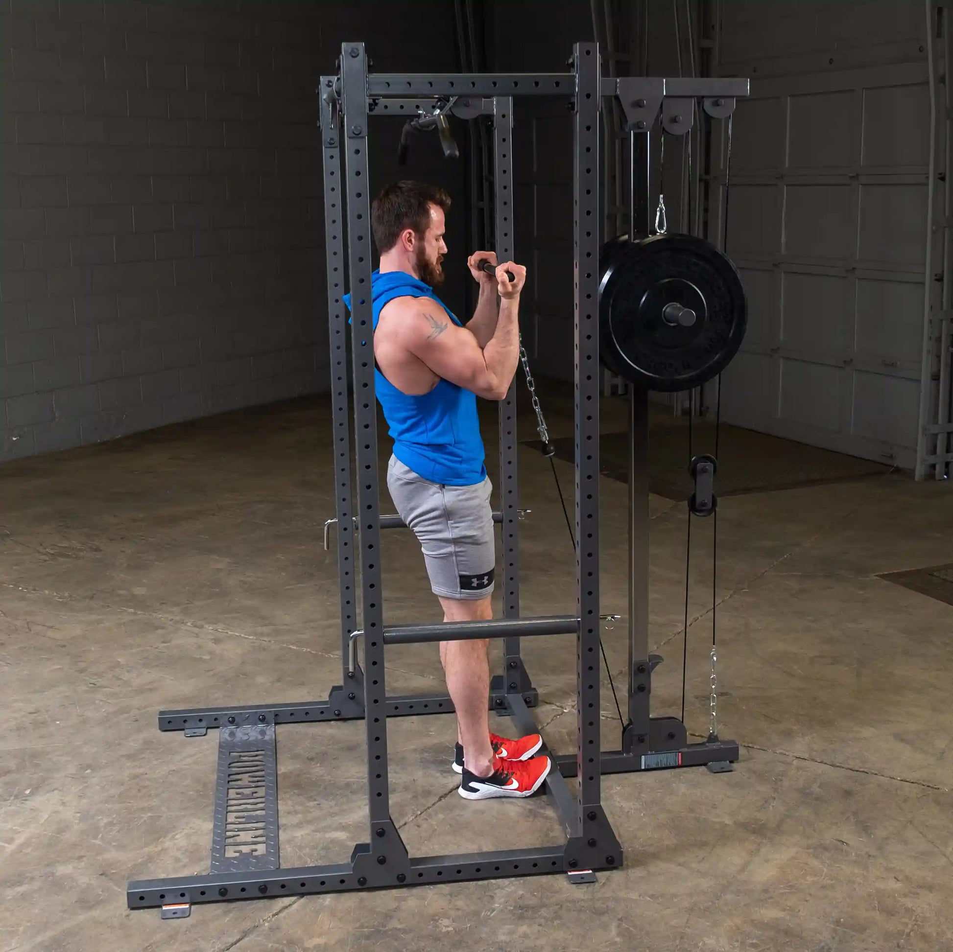 A muscular man wearing a blue tank top and gray shorts is performing an exercise using a cable machine in a gym. He is pulling a chain with weights, focusing on his biceps. The gym has a concrete floor and a simple background with a garage-style door.