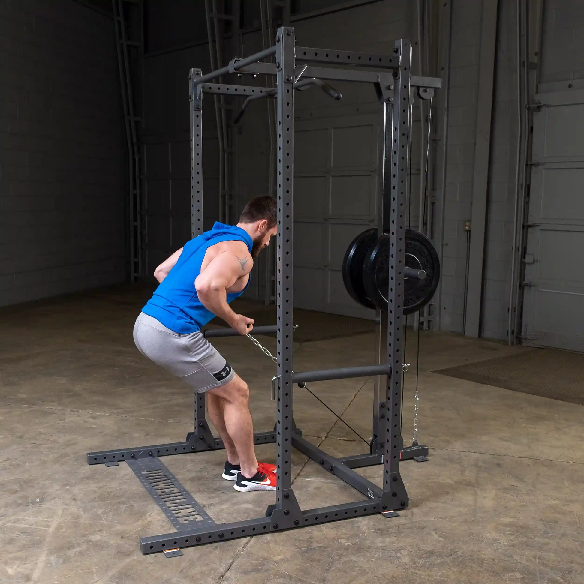 A man performing a bent-over row exercise using a cable machine in a gym setting. He is wearing a blue sleeveless hoodie and gray shorts, with red sneakers, demonstrating proper form.