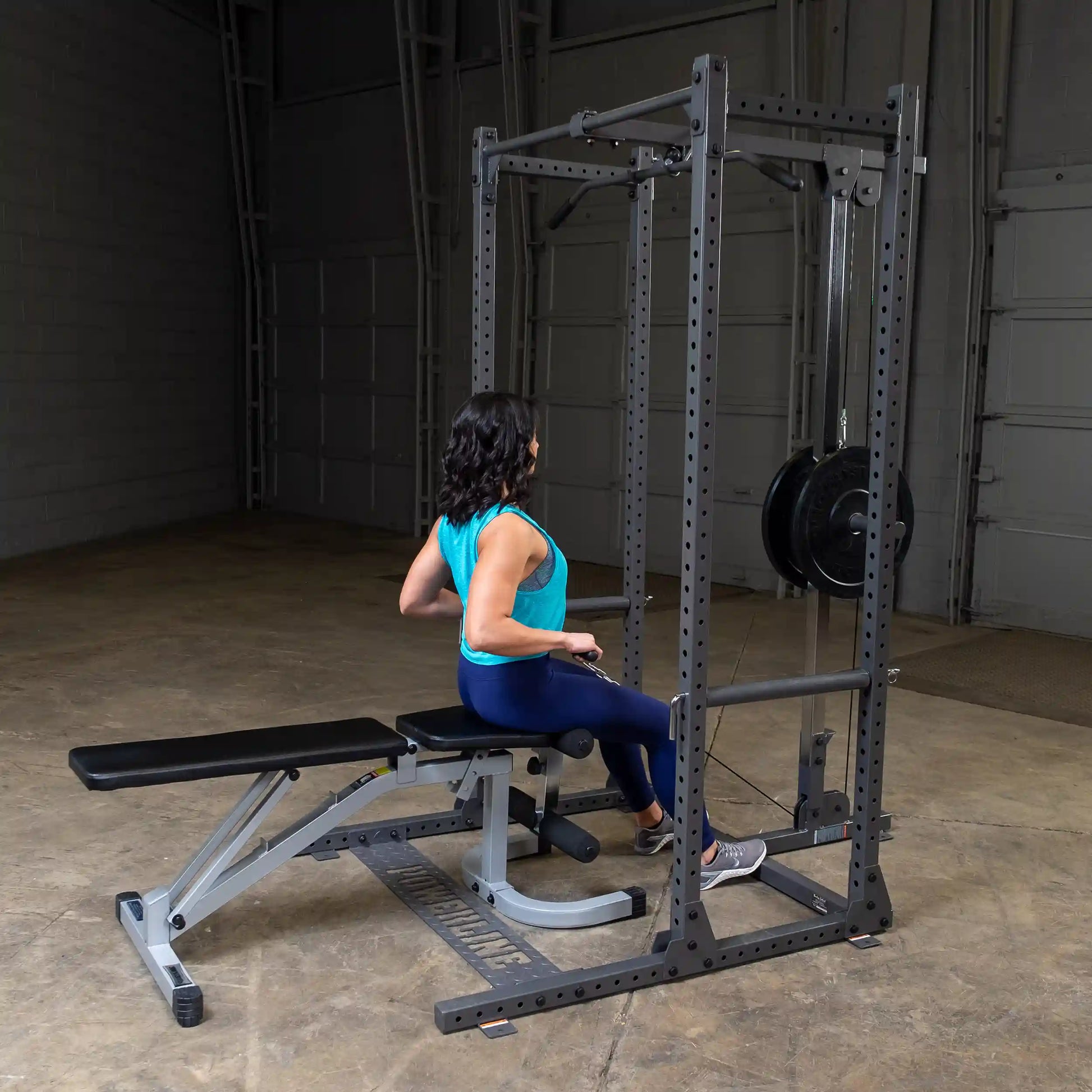 A woman in a turquoise tank top and blue leggings performing a seated row exercise using a gym machine in a spacious indoor setting.