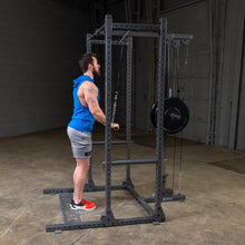 A man in a blue sleeveless hoodie and gray shorts performing a cable exercise in a gym, using a cable machine with weights attached.