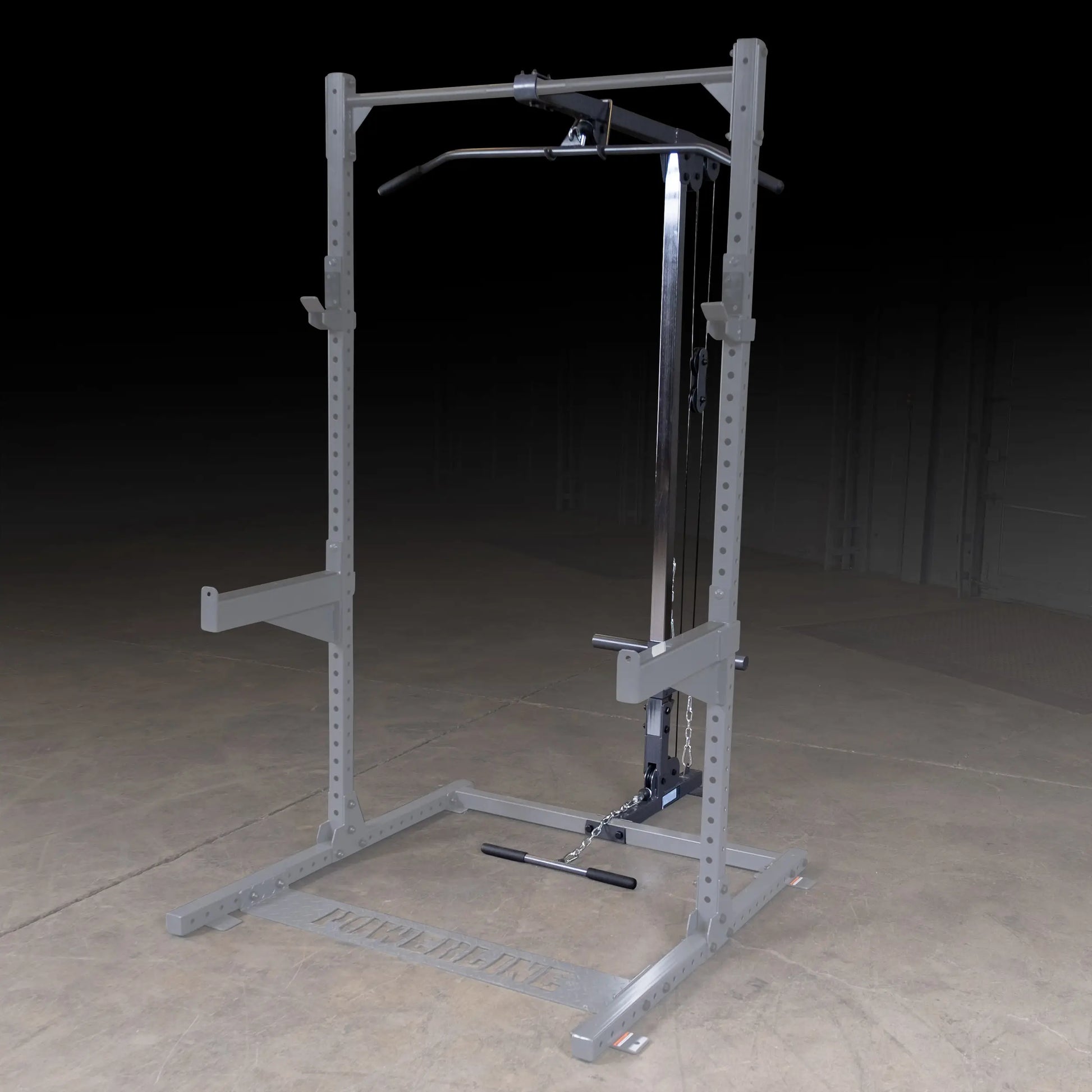 A gray weightlifting machine with a pull-up bar and adjustable cable system, set against a dark background on a concrete floor.