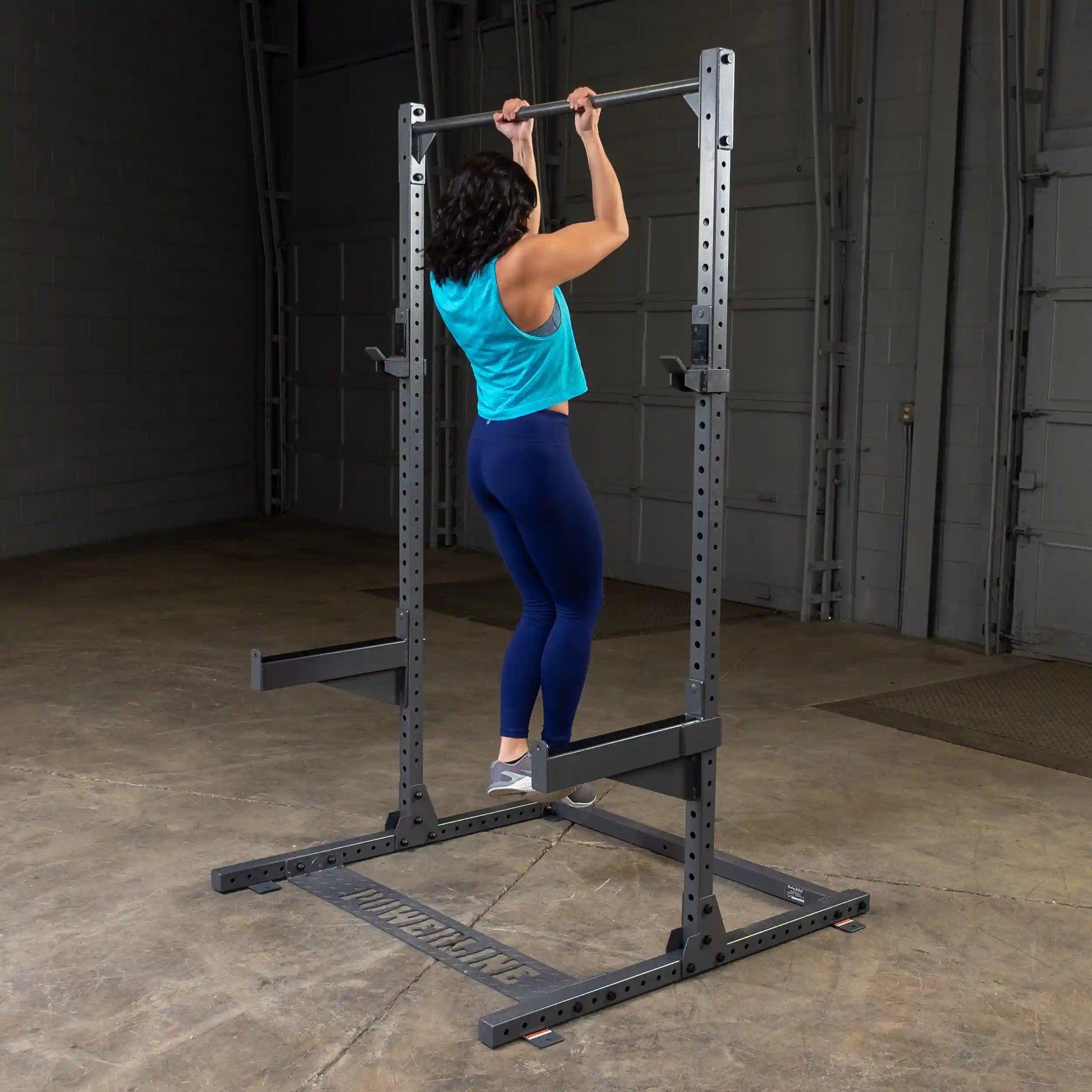 A person performing a pull-up on a squat rack in a gym setting, wearing a blue tank top and dark leggings, showcasing strength training exercises.