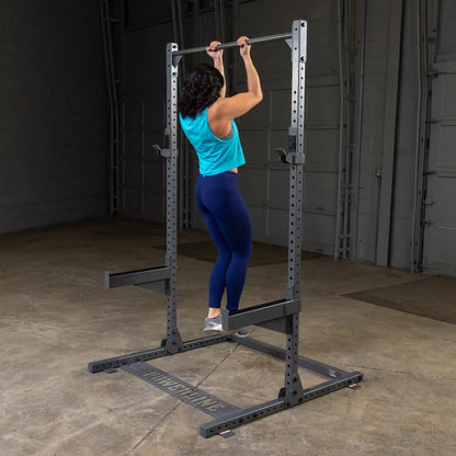 A person performing a pull-up on a squat rack in a gym setting, wearing a blue tank top and dark leggings, showcasing strength training exercises.