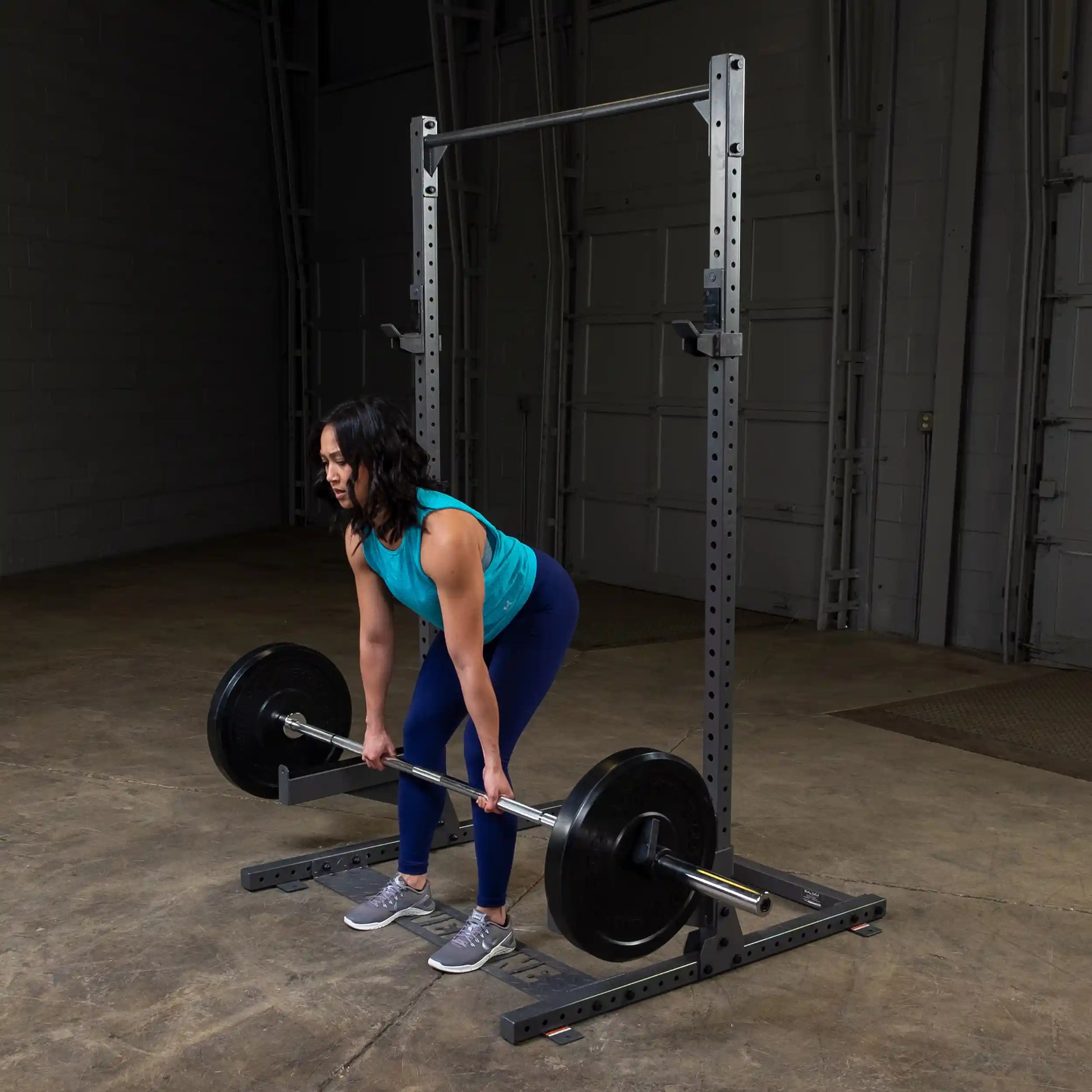 A woman with shoulder-length dark hair is performing a deadlift in a gym setting. She is wearing a blue tank top and navy leggings, focusing on lifting a barbell with weights. The background features gym equipment and a concrete floor.