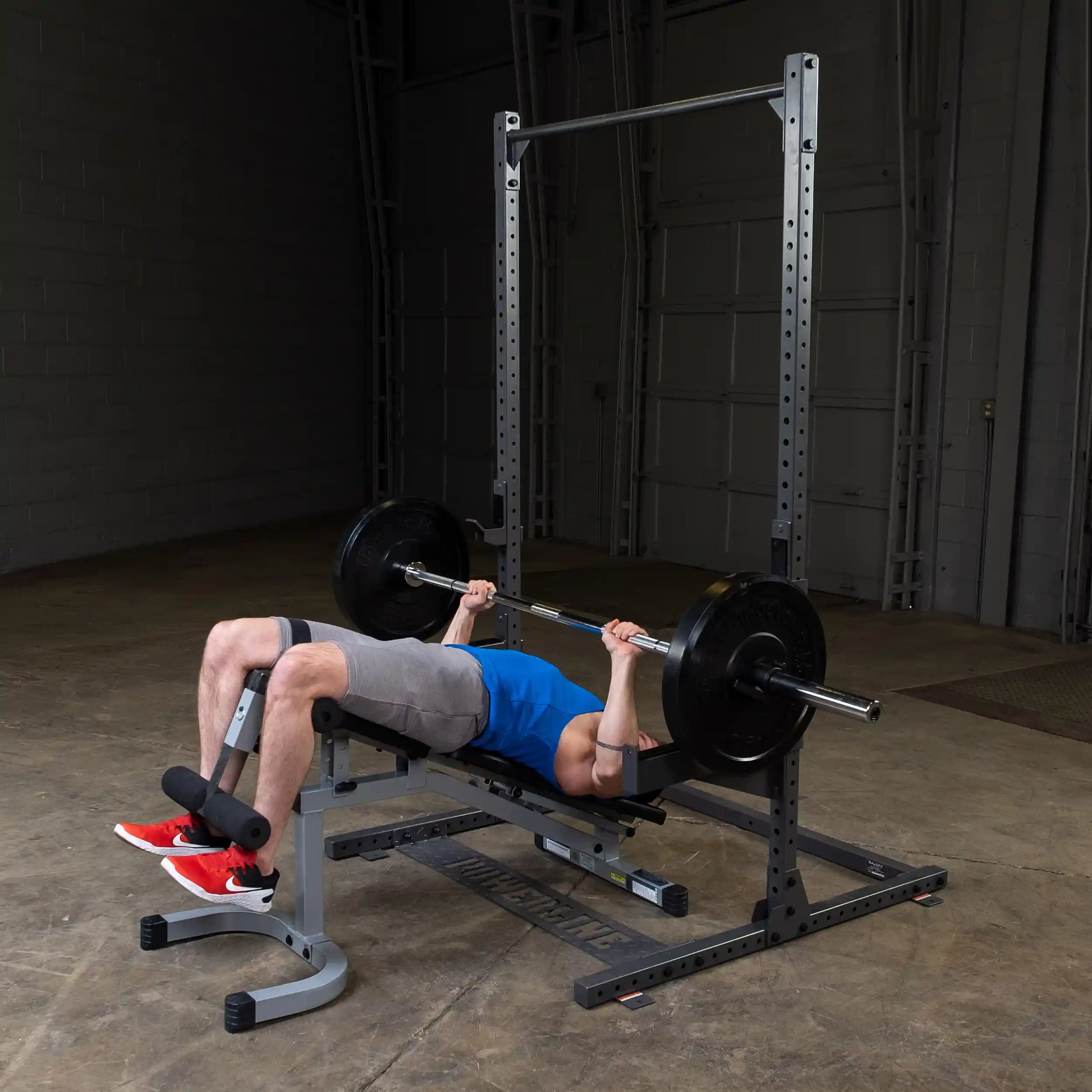 A person performing a bench press with a barbell and weights in a gym setting, wearing a blue shirt and gray shorts, with red sneakers, using a weight bench and rack.