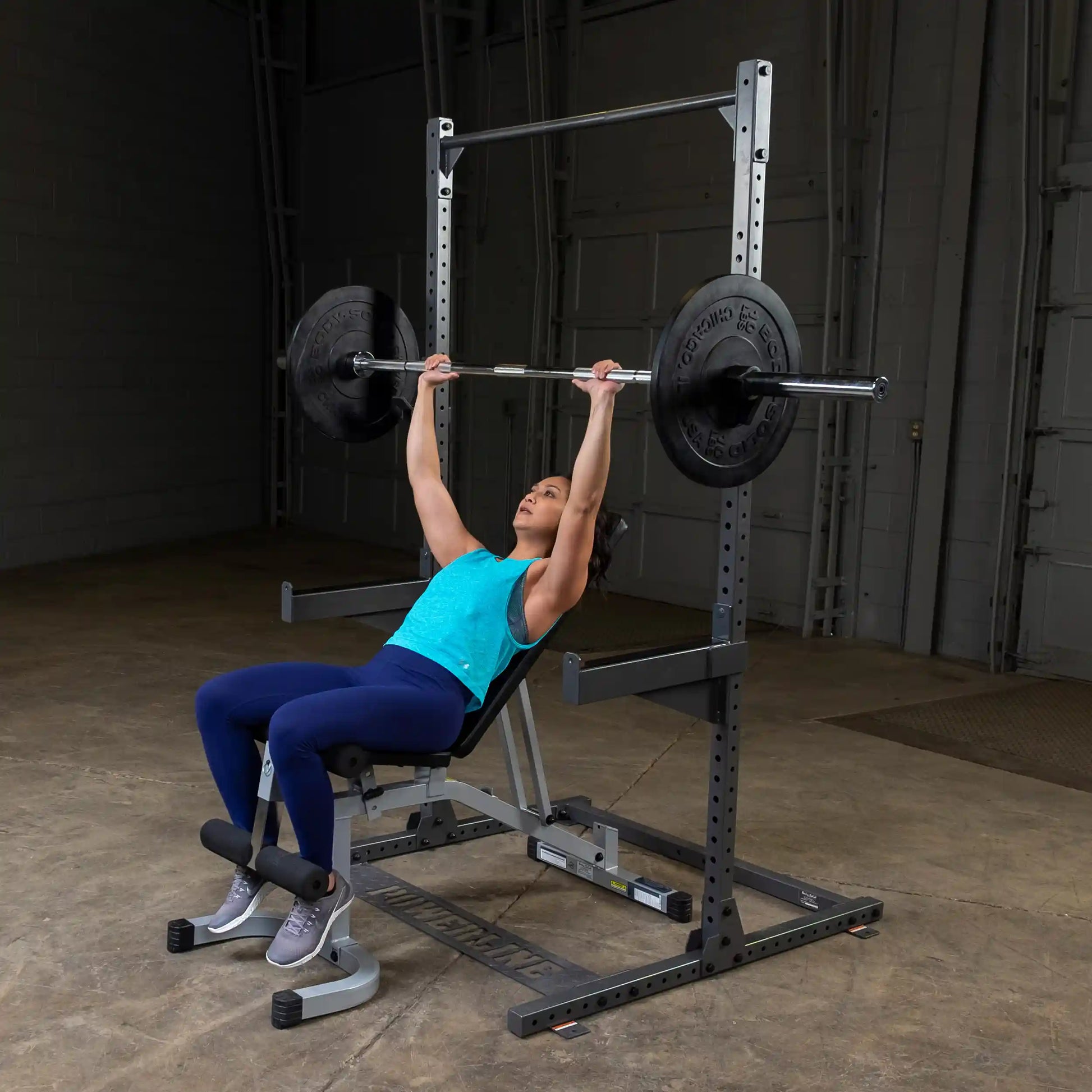 A woman performing a bench press exercise with a barbell and weights in a gym setting, wearing a blue tank top and navy leggings.