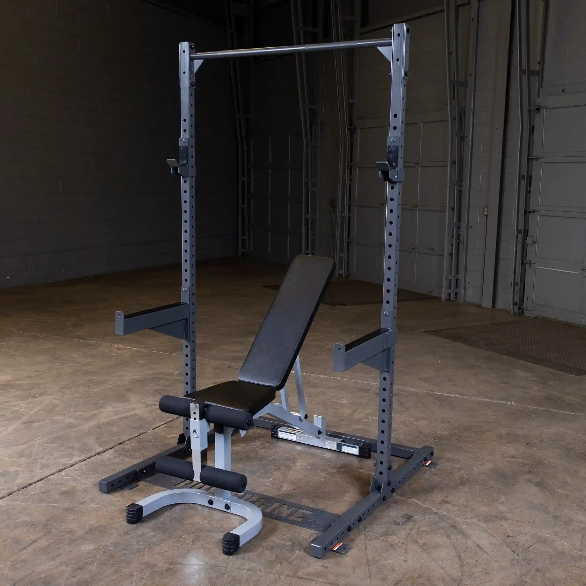 A gym setup featuring a weight bench and squat rack, positioned in a spacious, well-lit area with a concrete floor and gray walls.