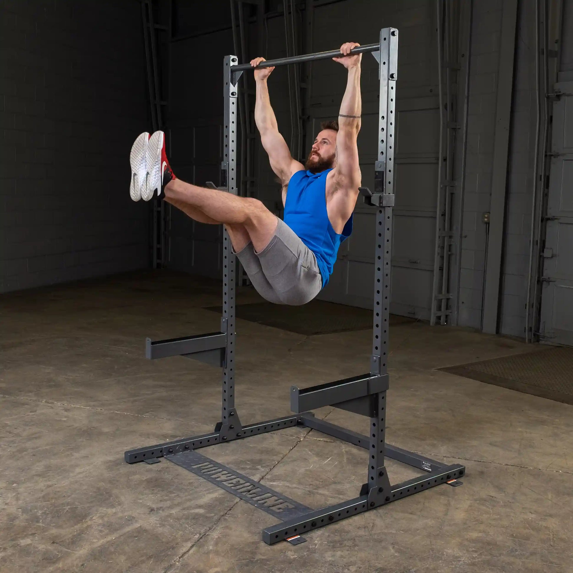 A man in a blue sleeveless shirt and gray shorts performs a hanging leg raise on a pull-up bar in a gym setting, showcasing strength and fitness.
