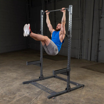 A man in a blue sleeveless shirt and gray shorts performs a hanging leg raise on a pull-up bar in a gym setting, showcasing strength and fitness.