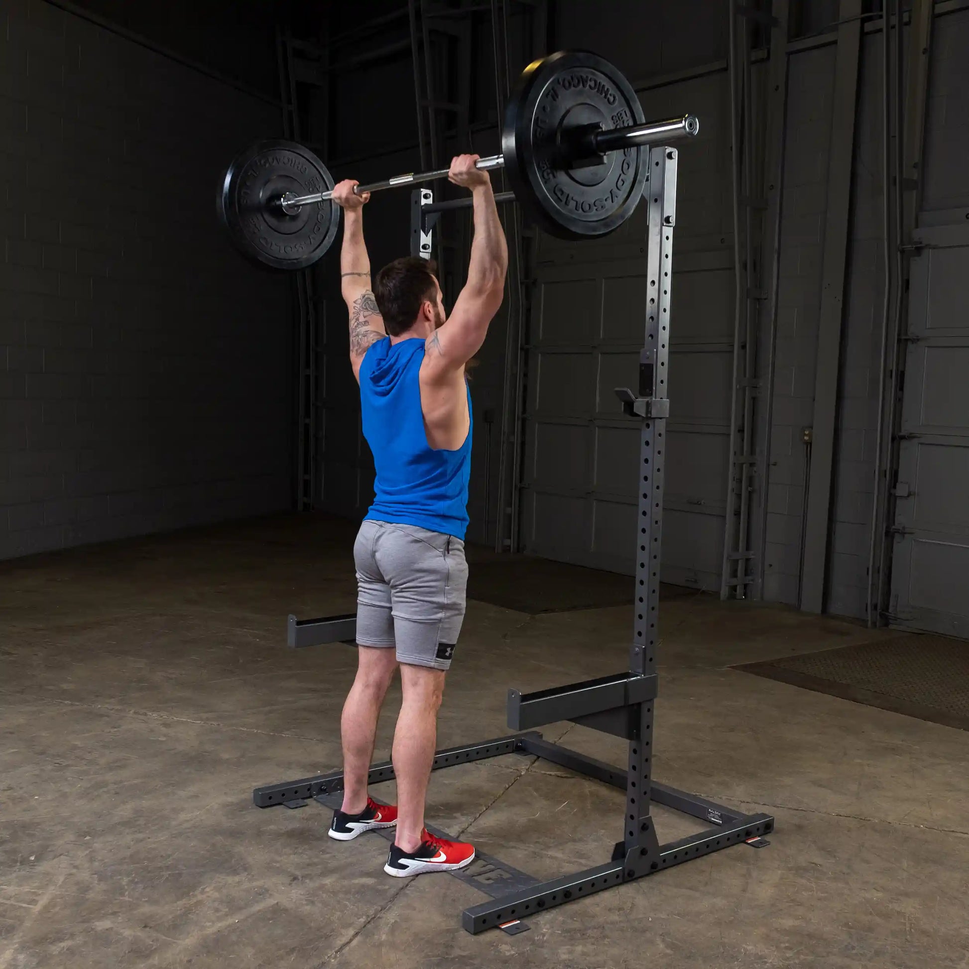 A person wearing a blue tank top and gray shorts is performing an overhead barbell lift in a gym setting. The individual is positioned in front of a squat rack, with weights on the barbell, showcasing strength training.