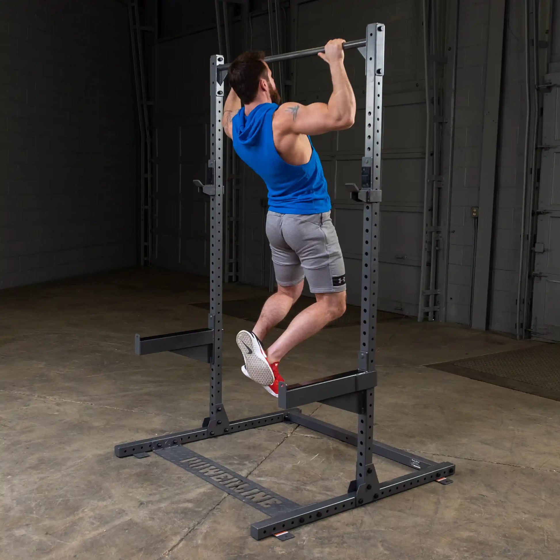 A person performing a pull-up on a strength training equipment in a gym setting, wearing a blue sleeveless hoodie and gray shorts.